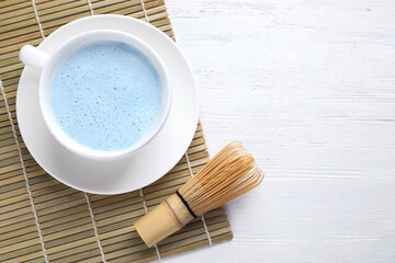 Fototapeta na wymiar Blue matcha latte, chasen and bamboo mat on white wooden table, flat lay