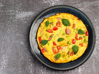 Omelet with tomatoes, sausage and bell pepper with herbs on a round plate on a dark background. Top view, flat lay
