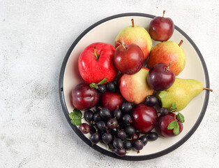 Raw fruits - apples, pears, plums and gooseberries on a round plate on a light gray background. Top view, flat lay