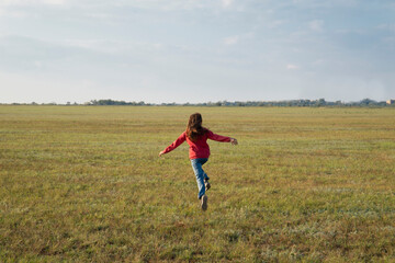 A girl with long hair in a red jacket runs across a green field