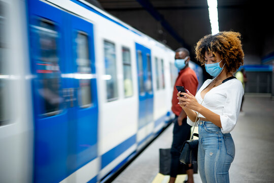 African American woman with face mask using smart phone waiting for the train on platform.