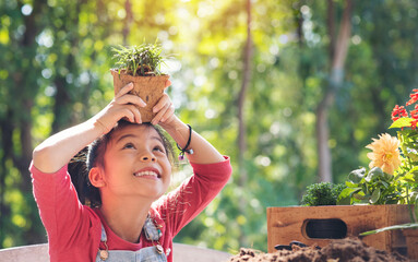 Cute child asian girl plays little gardener and holding flowerpot plant on head in spring garden,...