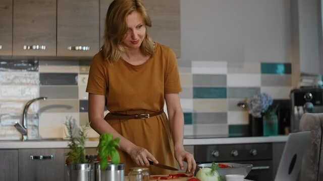 Young European woman cutting red pepper at table with computer in home kitchen rbbro. Beautiful female cuts fresh vegetable on desk and looks at laptop screen with smile, stands in light interior