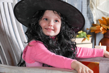 a girl in a witch's cap and wig sits on a chair in the halloween decorations, smiles and makes faces