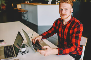 Portrait of young red hair man with modern cellular technology in hand sitting at coworking table desktop and looking at camera during break from laptop programming,Caucasian freelancer posing indoors