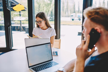 Fototapeta premium Young Caucasian woman in optical eyewear writing informative notes in education copybook during cooperation brainstorming meeting with blurred male colleague using smartphone application for calling