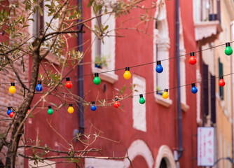 Wired colored bulbs hanging from one side of the street to the other over the street. Tree branches in front of colored bulbs. Old houses with many windows in the back