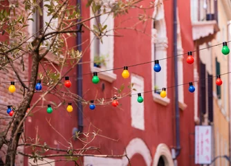 Fotobehang Wired colored bulbs hanging from one side of the street to the other over the street. Tree branches in front of colored bulbs. Old houses with many windows in the back © Vitalie