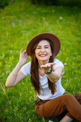 Young happy woman in a hat sits on a green lawn in a park. A girl of European appearance with a smile on her face on a bright sunny summer day