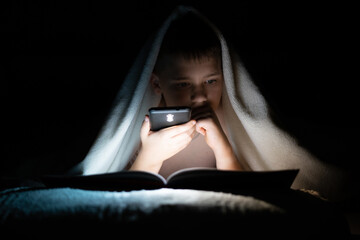 a boy lying in bed reading a book in the dark, illuminating with a flashlight from the phone