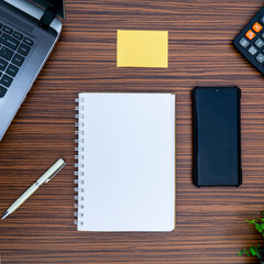 An office table working enviroment. Notepad, sticky note, pen plant, calculator and a lap top on a brown striped zebrawood design table top. Must have objects while working from home during Covid-19