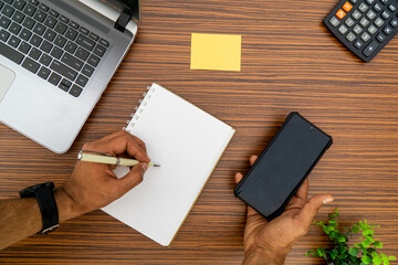 Writing on a notepad while holding a mobile phone working in an office environment. A lap top, a mobile, calculator and plant are also on display on this brown striped working table.