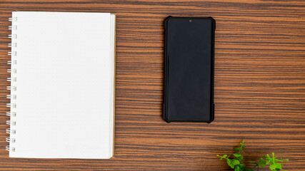 Personal note book and a black mobile phone with a glimpse of green plant on a striped brown office table