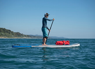 Active man rowing on SUP touring with waterproof bag at sunny day