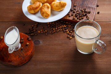 A mug of coffee with cream and a jug of cream and croissants on the table among the scattered coffee beans.