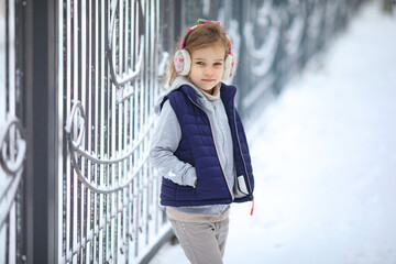 a little girl in warm headphones is standing at the fence in the winter in a snowy Park. hardening of children.