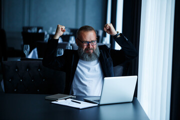 An adult male businessman is working on a new project and looking at the stock growth charts. Sits at the table by the large window. Looks at the laptop screen and smiles