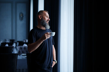 An adult man with a beard drinks coffee and looks out the window. Working day field recreation concept