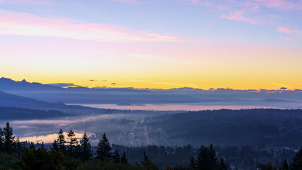 Mists rising over Burrard Inlet at Port Moody, BC, as orange glow of sunrise highlights distant mountains in silhouette