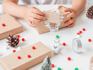Obraz na płótnie Canvas Kid wraps handmade Christmas presents in craft paper with colorful pompons and snowflake ribbons. Child prepares gifts for New Year celebration. Peaceful leisure activity before winter holidays.
