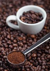 Fresh raw organic ground coffee powder in black steel scoop with white epresso cup on top of coffee beans.