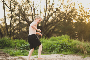 Boy dancing in sand with no shirt on. Vibrant sunlight coming through trees in background.