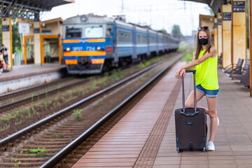 Safe travel. Caucasian teenager in black face mask with suitcase is waiting for train at the railway station.