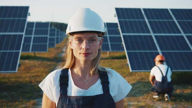 Portrait of nice-looking cheerful female blonde engineer in uniform standing by solar batteries. Woman worker cooperating with colleague on ecological solar farm.