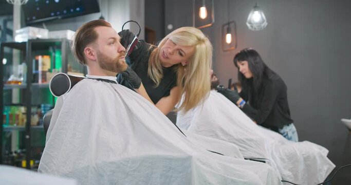 Young bearded man is sitting on the barber's chair, with cape, facing the mirror, female barber clippers his hair with hair clipper. Behind them, another female barber, who shaves with razor beard of