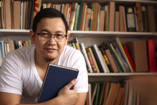 Portrait of cheerful Asian male librarian smiling, man standing against books in library