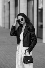 Obraz premium Young stylish woman in white overall, sneakers, black leather jacket and sunglasses holding small handbag and standing at city street. Fashion style outdoor portrait