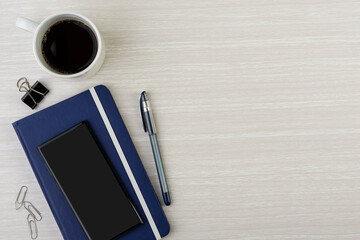 Note book with pen, smart phone, coffee mug and paper clips over wooden desk with room for text. Top view