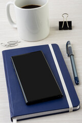 Note book with pen, smart phone, coffee mug and paper clips over wooden desk with room for text