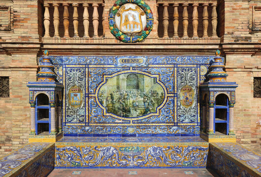 Image with the name of the spanish city of  Orense with a historical scene painted on ceramic tiles, with and the emblem shield above - seating benches in Spain Square in Seville