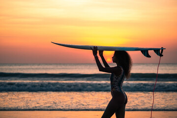 Portrait of woman surfer with beautiful body on the beach with surfboard at colorful sunset in Bali