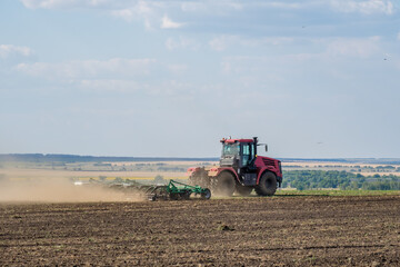 A red farm tractor in a cloud of dust cultivates the soil in the field with a cultivator after harvest. Summer sunny day. Fertile land. Modern agricultural machinery. Copy space. High quality photo