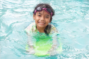 Fototapeta na wymiar Asian little girl wearing swimming goggles and holding a kickboard is practicing to float happily in the pool in the summer holiday.