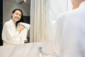 Beautiful young woman in white bathrobe with wet hair after taking a shower and washing her hair,happy asian teen girl standing in front of the mirror wiping hair with towel to dry in bathroom at home