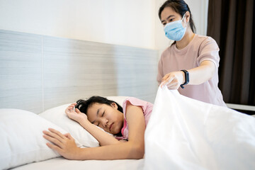 Sick female people sleeping on a bed in bedroom,asian mother in a mask cover child girl patient with blanket,putting up the blanket on her daughter at home,health care,love,family relations concept.