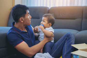 Asian single dad feeding milk to son relaxing in new house living room 
