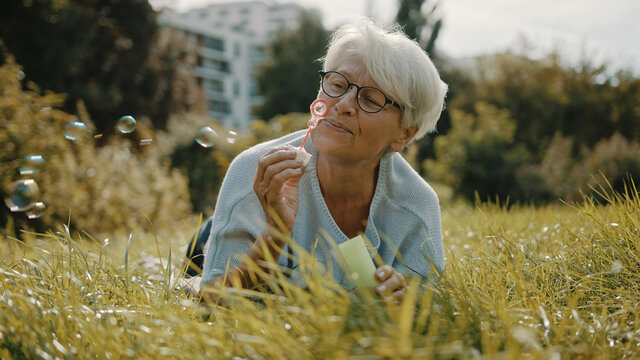 Retired senior woman enjoying freedom. Childish grandma blowing soap bubbles in the park. High quality photo