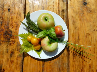 Still life: apples, cherry plums, grape leaves, hop cones and ears of a cereal plant lie on a white plate. The plate stands on a natural wooden background
