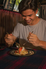 An action shoot of a young handsome man is enjoying his food using a fork and spoon to break a boiled egg on top of carbonara pasta meal in a restaurant