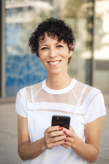 Fototapeta na wymiar Portrait of smiling mature woman with short black hair holding mobile phone in city