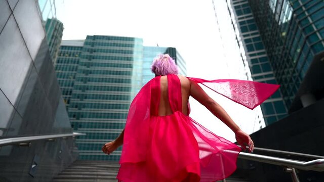 An unusual woman with pink hair whirls in the afternoon on the street against the background of the business center of the city. She's wearing a pink dress and a scarf.
