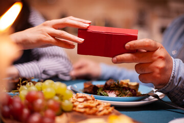 Man giving wife gift box and having romantic dinner together. Cheerful man dining with woman at home, enjoying the meal, celebrating their anniversary at candle lights.