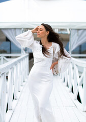young caucasian female with a thin figure in a white dress and white jacket touching her brown hairs, on the white boardwalk near the railings. summer house at the background