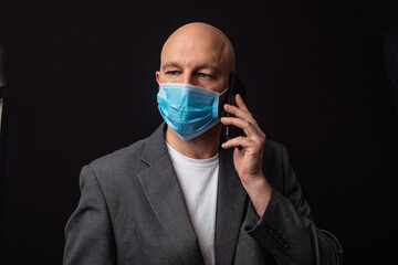 Portrait of a bald businessman in white t-shirt and grey suit on dark background, talking on black smart phone and wearing blue protective face mark during COVID 19 flue outbreak.