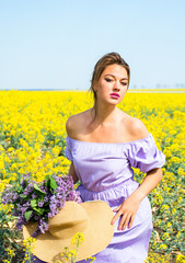 young caucasian female with hat and lilac bouquet in hand in magenta dress is sitting in the yellow field filowers and enjoy the warmth of the sun
