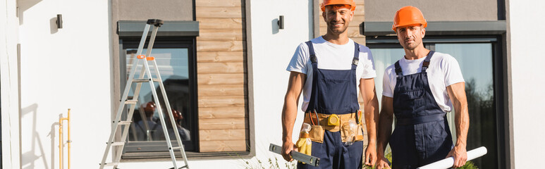 Horizontal crop of builders in uniform holding tools and blueprint near building